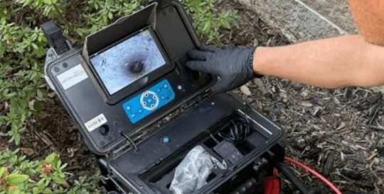 A technician is examining a screen displaying a pipe interior using a specialized camera system in a black carrying case.