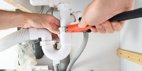 A plumber is using a wrench to work on pipes under a sink.
