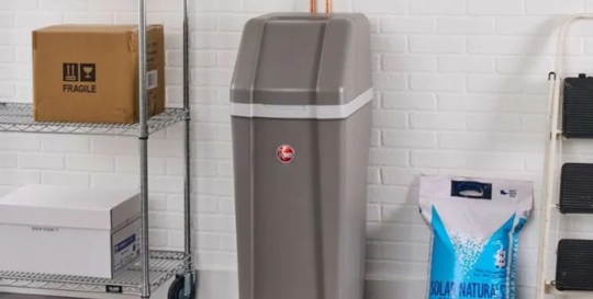 A water softener stands in a storage room with shelves, boxes, and a bag of salt.