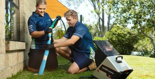 Two uniformed workers, one kneeling, insert a hose into a pipe on a lawn.