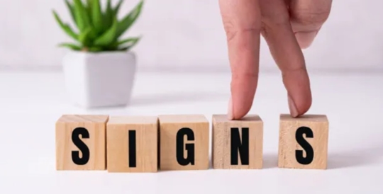 The image shows wooden blocks spelling out the word "SIGNS," with fingers walking on top of the blocks