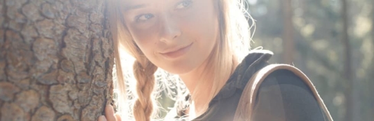 A woman with blonde braids smiles, leaning against a tree trunk.