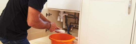 A plumber is shown repairing pipes under a sink.