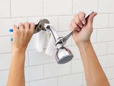 A person is using two wrenches to remove a showerhead from a tiled wall, with a cloth protecting the fixture.