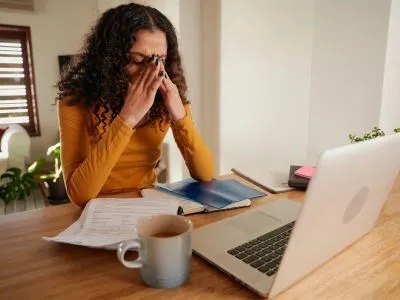 A woman sits at a desk with a laptop and documents, appearing stressed or overwhelmed as she rubs her eyes.