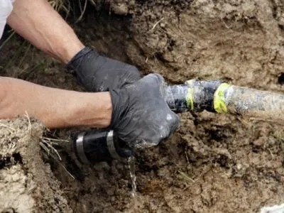 A person wearing black gloves is repairing a leaking pipe in a ditch.