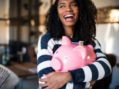 A happy woman hugs a pink piggy bank with money sticking out of it.