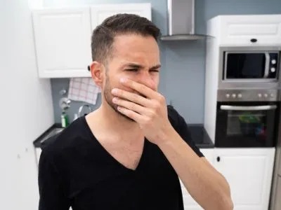 A man in a kitchen holds his hand over his nose, reacting to a bad smell.