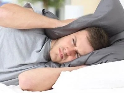 A man in bed looks annoyed as he covers his ears with a pillow.