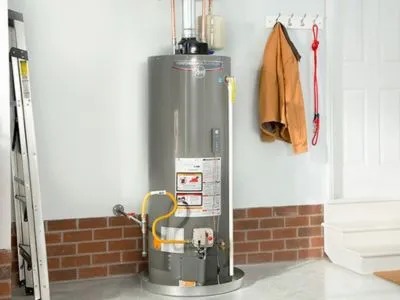 A gas water heater stands against a brick-trimmed wall in a garage-like setting.