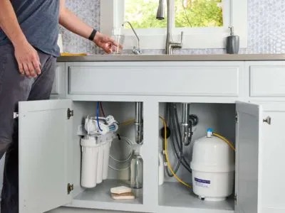 A man opens the cabinet under a kitchen sink to reveal a water filtration system.