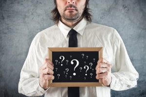 A businessman is holding a small chalkboard covered with question marks.